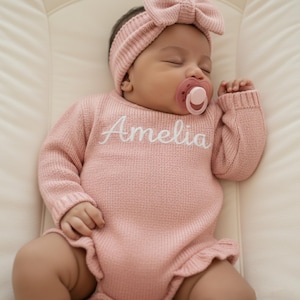 Peut inclure: Un bébé endormi portant un body rose en tricot avec le nom "Amelia" brodé en blanc. Le bébé porte également un bandeau assorti avec un nœud et une sucette. Le body a un ourlet à volants.