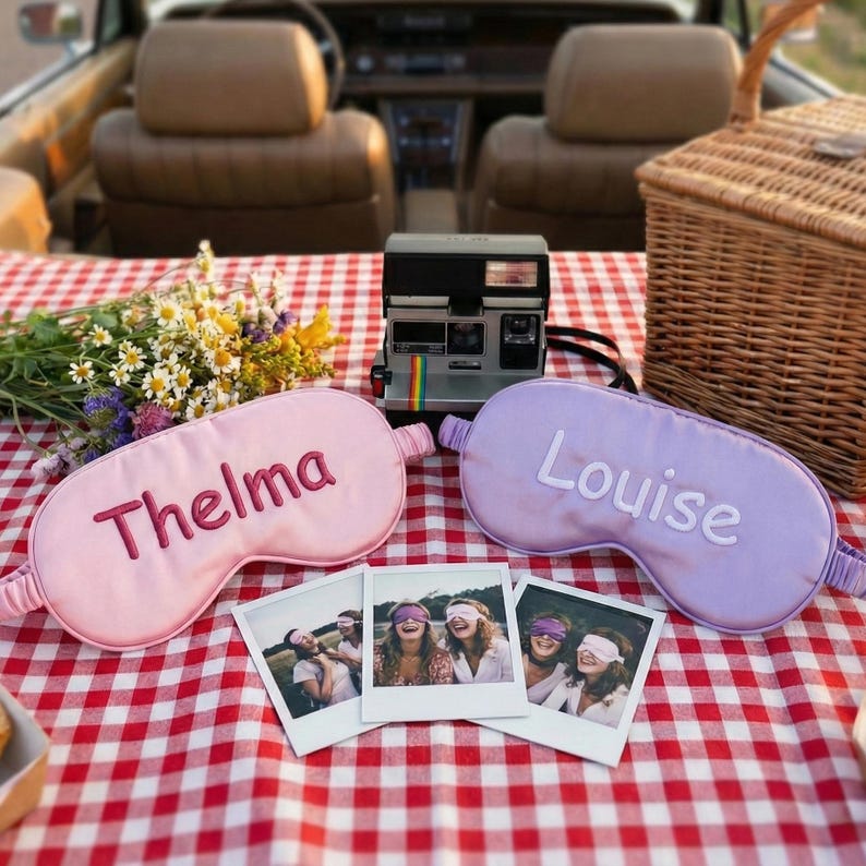 May include: A picnic scene with a red and white checkered tablecloth. Two sleep masks, one pink with "Thelma" embroidered and one lavender with "Louise". A vintage Polaroid camera and three instant photos are also on the table.