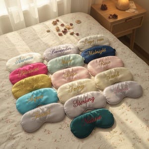 May include: A collection of satin sleep masks in various colors, including pink, blue, and yellow, each embroidered with a different name. The masks are arranged on a floral patterned bedspread. The names include "Aurora", "Blossom", and "Sunshine".