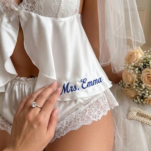 Pu&ograve; includere: Completo intimo da sposa in raso bianco con finiture in pizzo. Il set comprende un top e pantaloncini, con la scritta ricamata "Mrs. Emma" in blu. &Egrave; visibile un anello di diamanti.