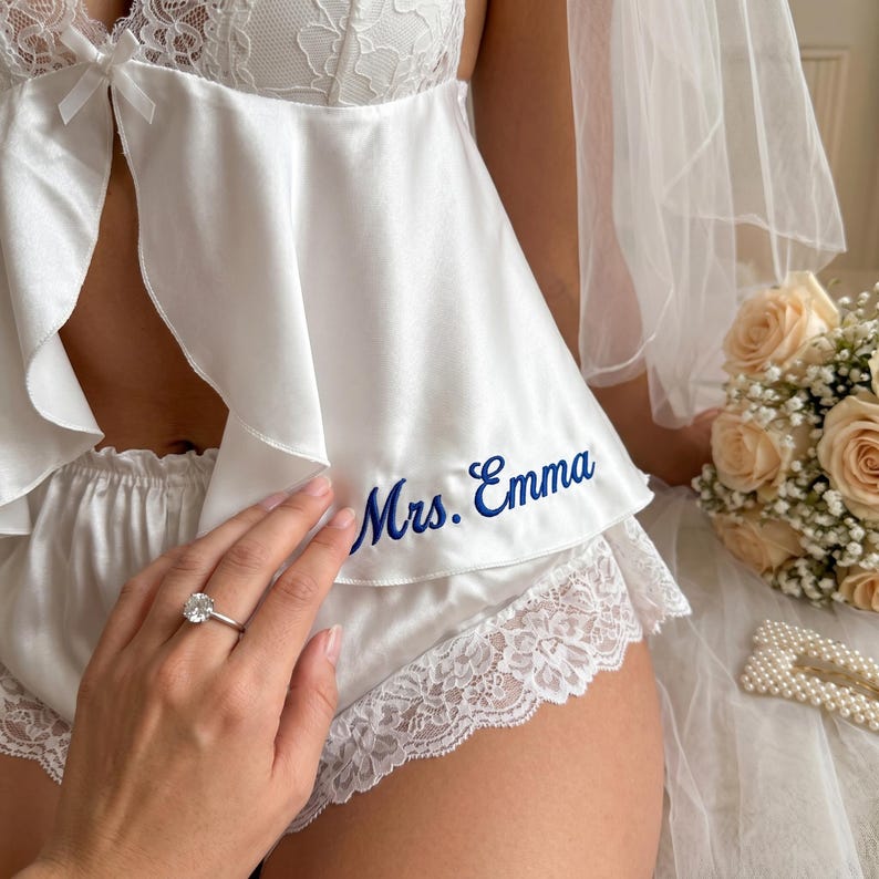 Pu&ograve; includere: Completo intimo da sposa in raso bianco con finiture in pizzo. Il set comprende un top e pantaloncini, con la scritta ricamata "Mrs. Emma" in blu. &Egrave; visibile un anello di diamanti.