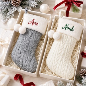 May include: Two Christmas stockings in individual gift boxes. One stocking is grey cable knit with a white faux fur cuff embroidered with the name Ava in red. The other is cream cable knit with a white faux fur cuff embroidered with the name Noah in green. Both feature two white pom-poms.