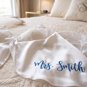 Pu&ograve; includere: Una vestaglia di raso bianco con la scritta ricamata "Mrs. Smith" in blu &egrave; appoggiata su un copriletto trapuntato color crema. Un reggiseno di pizzo bianco e un velo sono anch'essi presenti, suggerendo un tema nuziale.