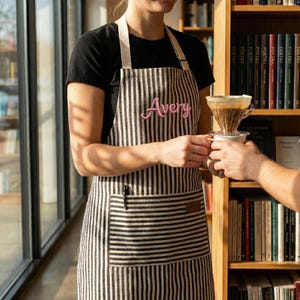 Puede incluir: Un delantal a rayas blancas y negras con el nombre "Avery" bordado en rosa. El delantal lo lleva una persona con una camisa negra, que está entregando una taza de café a otra persona. Un filtro de café está encima de la taza.