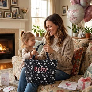 May include: A black tote bag featuring a white floral and celestial design, personalised with the name "Ruby" in pink. The bag is held by a woman and a child. The scene includes balloons, a Mother's Day card, and a gift box.