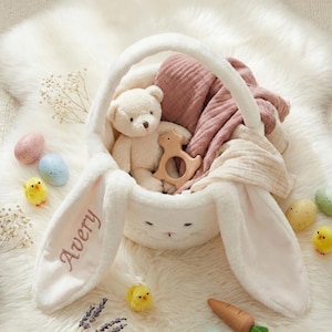 May include: A white plush bunny basket with long ears, embroidered with the name "Avery". Inside are a teddy bear, wooden toy, and pink blanket. Easter eggs and chicks surround the basket, creating a festive scene.