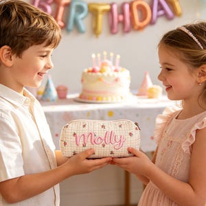 May include: A beige pencil case with a floral pattern and the name "Molly" embroidered in pink. The case is being held by two children, with a birthday cake and balloons in the background.