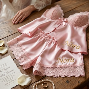 Pu&ograve; includere: Un completo pigiama in raso rosa chiaro con rifiniture in pizzo, composto da un top a camisole e pantaloncini. Il nome "Nora" &egrave; ricamato in oro su entrambi i pezzi. Sono visibili anche un invito di nozze, perle e un anello.