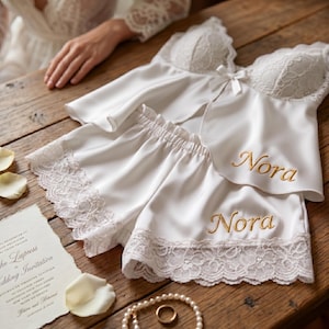 Pu&ograve; includere: Completo pigiama in raso bianco con bordi in pizzo, composto da top a camisole e pantaloncini. Il nome "Nora" &egrave; ricamato in oro su entrambi i capi. Sono visibili anche un invito di nozze e una collana di perle.