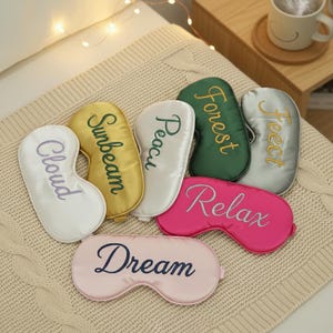 May include: A collection of satin sleep masks in various colors, including white, gold, green, silver, pink, and light pink. Each mask is embroidered with a different word: "Cloud", "Sunbeam", "Peace", "Forest", "Feet", "Relax", and "Dream".