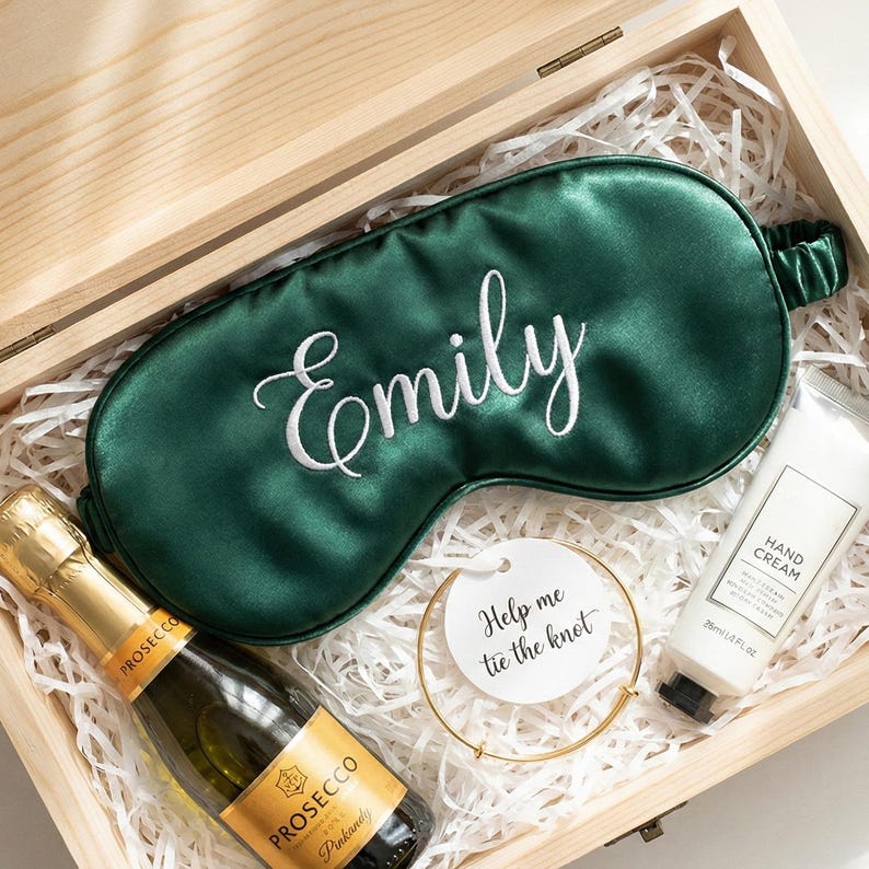 May include: A wooden gift box filled with white paper, a green satin sleep mask embroidered with "Emily", a bottle of Prosecco, a gold bracelet with a tag that says "Help me tie the knot", and a tube of hand cream.