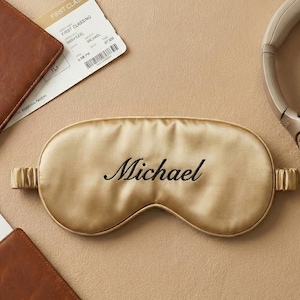 May include: A gold-colored satin sleep mask embroidered with the name "Michael" in black script. The mask is surrounded by travel accessories, including a boarding pass, headphones, and a leather wallet.