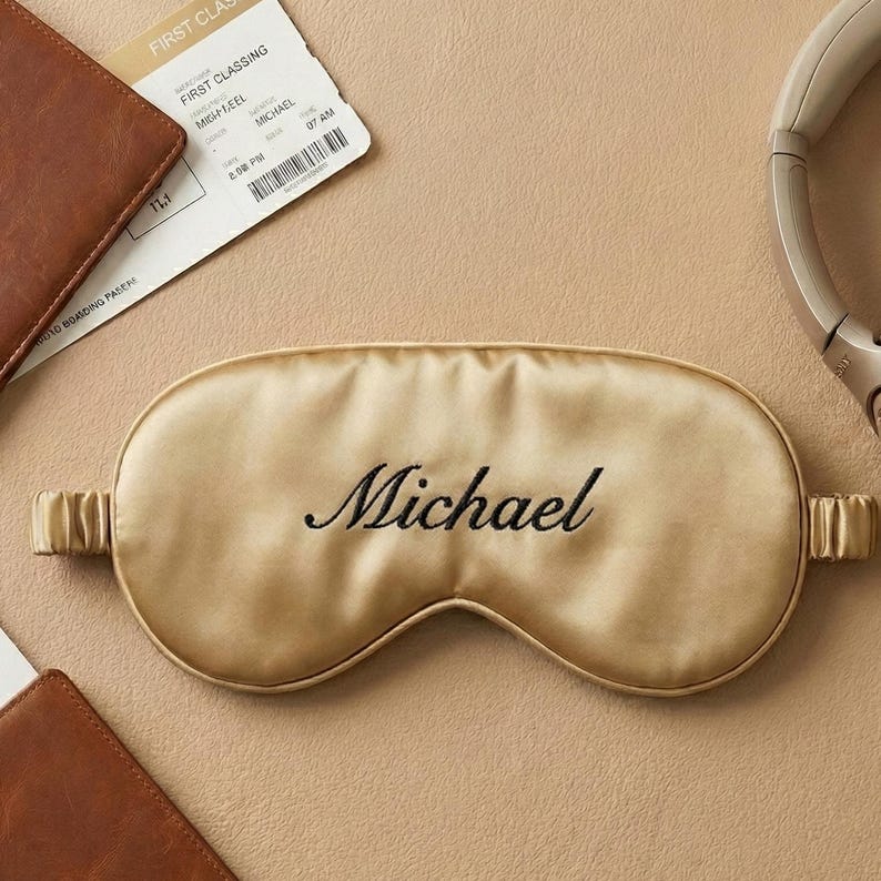 May include: A gold-colored satin sleep mask embroidered with the name "Michael" in black script. The mask is surrounded by travel accessories, including a boarding pass, headphones, and a leather wallet.