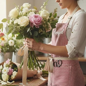 May include: A florist arranging a bouquet of white roses, a pink peony, and greenery. The bouquet is tied with a cream-colored ribbon. The florist wears a red and white striped apron with the name "Angel" on it.