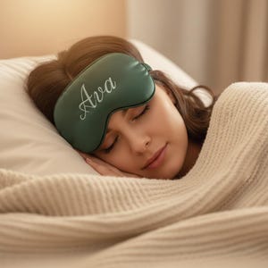 May include: A person wearing a dark green sleep mask with the name "Ava" in white script. The person is lying on a white pillow, covered with a cream-colored blanket. The scene is softly lit.