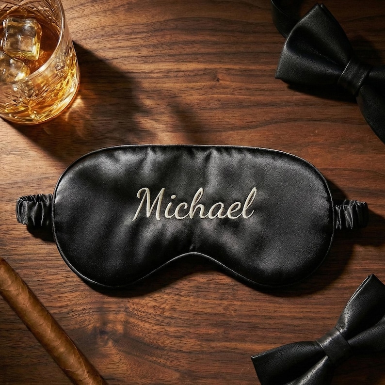 May include: Black satin sleep mask embroidered with the name "Michael" in white script. The mask is surrounded by a glass of whiskey with ice, a cigar, and a black bow tie, all on a wooden surface.