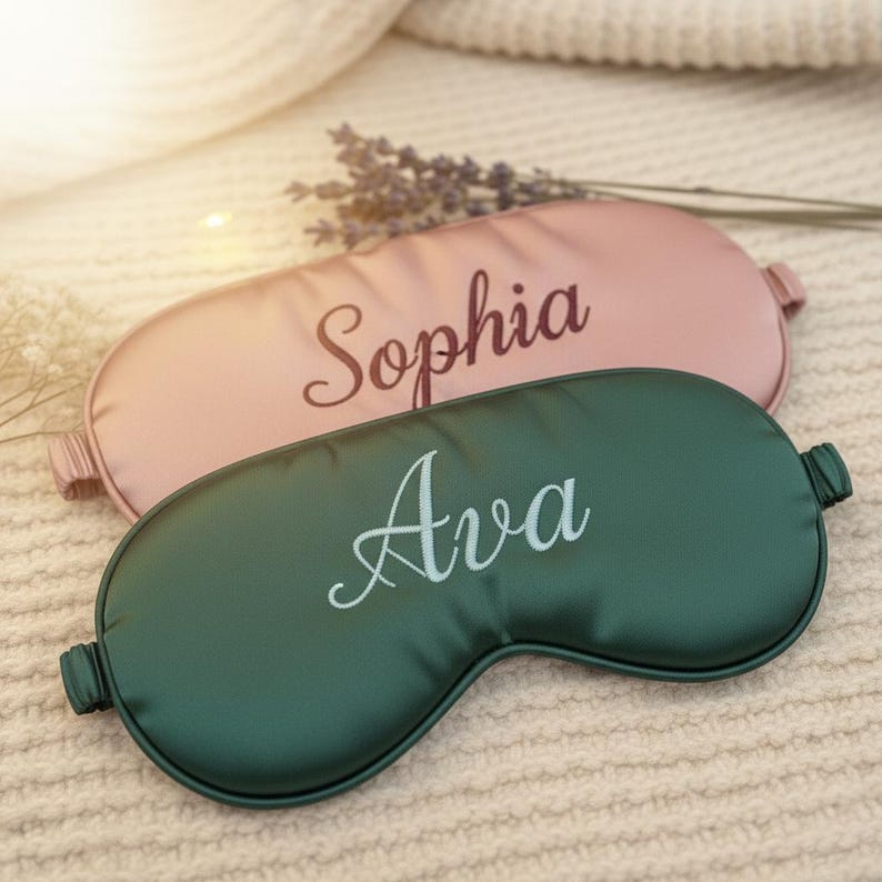 May include: Two personalized sleep masks. One is a soft pink color with the name "Sophia" embroidered in a cursive font. The other is a deep green color with the name "Ava" embroidered in a similar font. Both masks are made of a smooth, satin-like material.