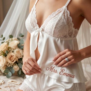 Pu&ograve; includere: Completo intimo in raso bianco con finiture in pizzo. Il top presenta uno scollo in pizzo e un piccolo fiocco. Anche i pantaloncini sono rifiniti in pizzo. Il testo "Mrs. Emma" &egrave; ricamato in corsivo sui pantaloncini.