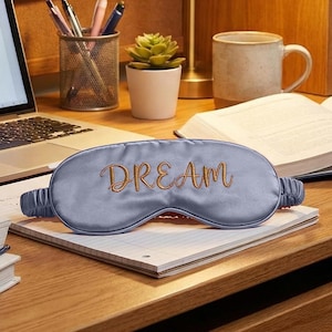 May include: A light blue satin sleep mask with the word "DREAM" embroidered in gold script. The mask is resting on a notepad on a wooden desk, with a laptop, mug, and plant in the background.