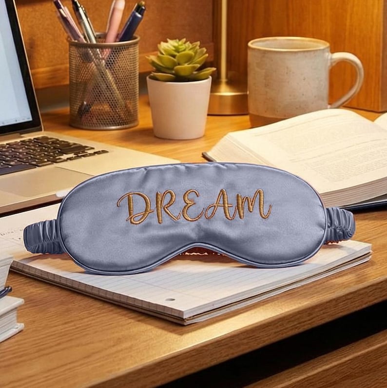 May include: A light blue satin sleep mask with the word "DREAM" embroidered in gold script. The mask is resting on a notepad on a wooden desk, with a laptop, mug, and plant in the background. The sleep mask is designed to block out light.