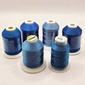 May include: Six spools of blue thread in varying shades, arranged on a white surface. The thread is wound around white plastic spools. Some spools have labels with numbers. The threads range from light to dark blue.