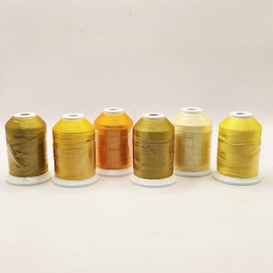 May include: Six spools of thread in various shades of gold and yellow, displayed on white plastic bases. The colors range from a deep olive to a pale cream, with one spool in a bright orange hue. The thread is tightly wound.