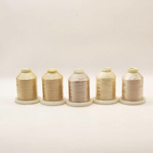 May include: Five spools of thread in varying shades of beige and gold. Each spool is cylindrical, with a white base and a yellow label on top. The threads appear to be tightly wound, creating a smooth, textured surface.