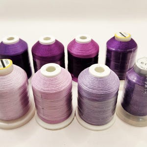 May include: Eight spools of thread in various shades of purple and lavender. The spools are arranged in two rows, with the colors ranging from light lavender to deep purple. Each spool is on a white base.