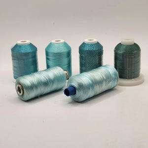 May include: Six spools of thread in various shades of teal, turquoise, and seafoam green. Four spools stand upright on white plastic bases, while two spools lie horizontally in the foreground. The thread has a smooth, slightly shiny finish.