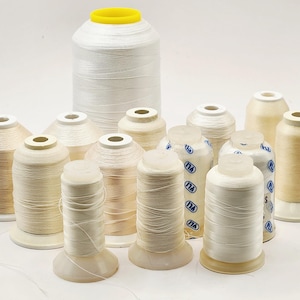 May include: A collection of thirteen spools of thread in various shades of white, cream, and beige. The spools are arranged in a group, with one large white spool featuring a yellow top cap positioned at the back and smaller spools in front.