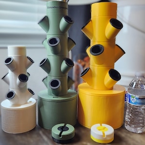 May include: Three vertical hydroponic towers in white, sage green, and bright yellow, each featuring multiple angled planting ports with black inserts. A clear plastic water bottle stands to the right, and two small circular base components sit in front.