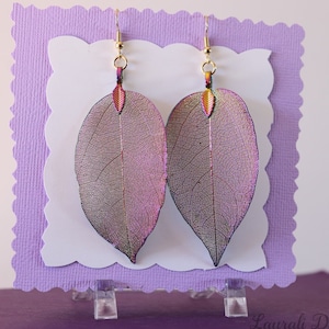 May include: A pair of iridescent leaf earrings with a pink and purple hue. The earrings are made from real leaves that have been preserved and coated with a clear resin. The earrings are hanging from gold-colored hooks.