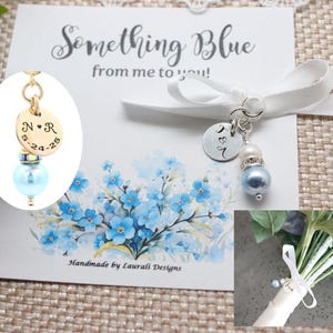 May include: A wedding keepsake with a light blue bead and a gold charm engraved with initials and a date. The charm is attached to a card that says "Something Blue from me to you!" with a floral design. A small bouquet is also shown.