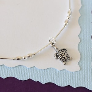 Può includere: Un braccialetto per caviglia in argento con un piccolo ciondolo a forma di tartaruga e perline d'argento. Il braccialetto è su uno sfondo bianco con un bordo blu e viola.