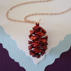 May include: A red metallic pine cone pendant necklace on a rose gold chain. The pendant is detailed and realistic.