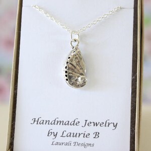 May include: A silver necklace with a pendant shaped like a seashell. The pendant is detailed with a textured surface and a small hole at the top. The necklace is displayed on a white card with the text "Handmade Jewelry by Laurie B Laurali Designs".