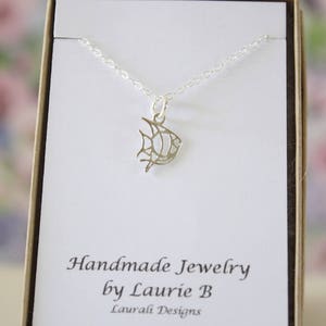 Op de afbeelding: Een zilveren ketting met een delicaat visje als hanger. De ketting wordt gepresenteerd op een witte kaart met de tekst "Handmade Jewelry by Laurie B Lauvali Designs".