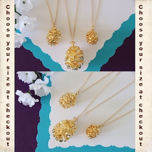 May include: Three gold pine cone necklaces of different sizes. The necklaces are on a white background with a turquoise border.