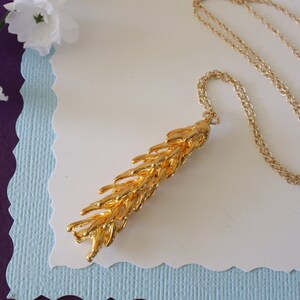 May include: A gold chain necklace with a long, gold pendant that resembles a wheat stalk.