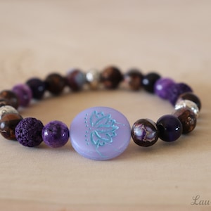 Può includere: Braccialetto con perline viola, marroni e argentate con un ciondolo lavanda con un designo a forma di fiore di loto blu.