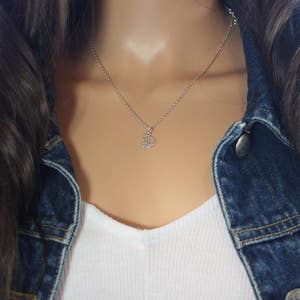 Op de afbeelding: Een zilveren ketting met een klein, delicaat hangertje. De ketting wordt gedragen door een persoon met lang, donker haar en een spijkerjack.