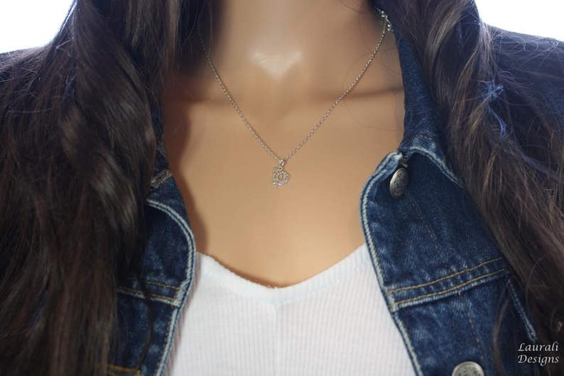 Op de afbeelding: Een zilveren ketting met een klein, delicaat hangertje. De ketting wordt gedragen door een persoon met lang, donker haar en een spijkerjack.