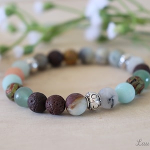 May include: A bracelet made with various natural stones, including green aventurine, brown lava rock, and grey agate. The bracelet has a silver charm with a decorative design.