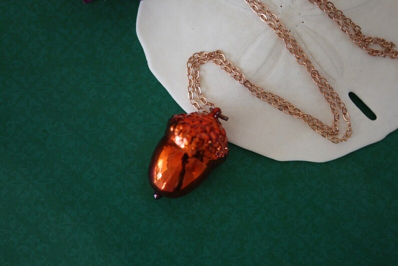 May include: A copper-colored acorn pendant necklace with a delicate chain. The acorn is detailed with a textured surface and a shiny finish.