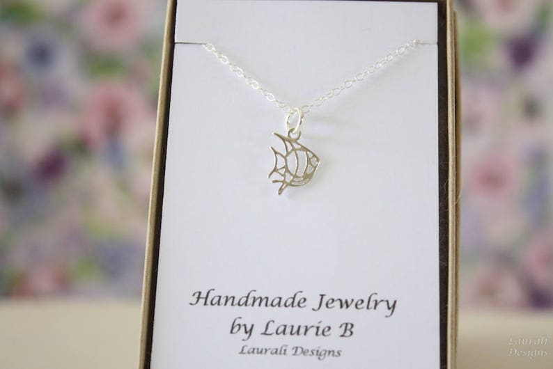 Op de afbeelding: Een zilveren ketting met een delicaat visje als hanger. De ketting wordt gepresenteerd op een witte kaart met de tekst "Handmade Jewelry by Laurie B Lauvali Designs".