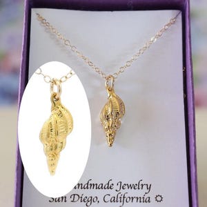 May include: Gold seashell necklace with a delicate chain. The pendant is a detailed, three-dimensional seashell. The necklace is displayed in a purple velvet box with a white card that reads "Handmade Jewelry San Diego, California".