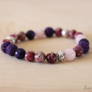 May include: A beaded bracelet featuring a variety of stone beads in shades of purple, pink, and maroon. The bracelet includes smooth, round beads, textured beads, and silver-colored accent beads. The bracelet is displayed on a light-colored wooden surface.