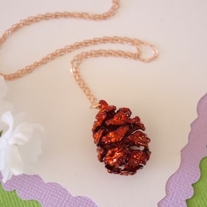 May include: A rose gold chain necklace with a red metallic pine cone pendant.