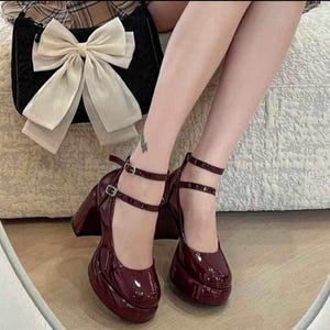 May include: A pair of dark burgundy patent leather platform heels with double ankle straps and silver-toned buckles, paired with a black handbag featuring a large, cream-colored fabric bow on the front and a plaid pattern on the top edge.