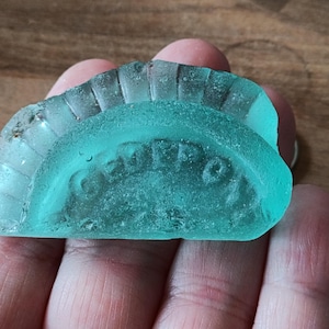 Puede incluir: Un trozo de vidrio marino en forma de semicírculo, de color verde mar, con la palabra "CORPO" en relieve. El borde superior tiene un diseño estriado. El vidrio es translúcido y tiene un aspecto desgastado.